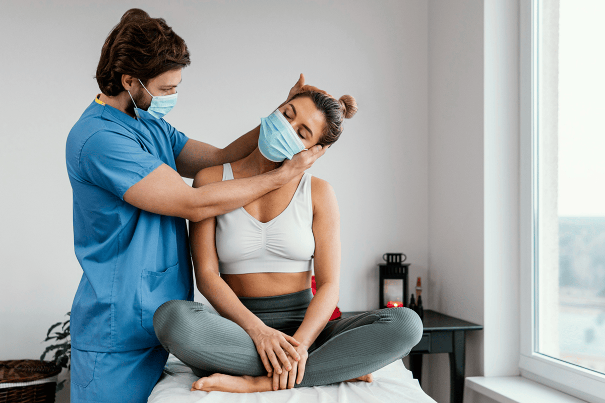 A young woman sitting on medical table, 
            wearing a facemask and workout clothes, is stretched by a physical therapist, 
            also wearing a mask